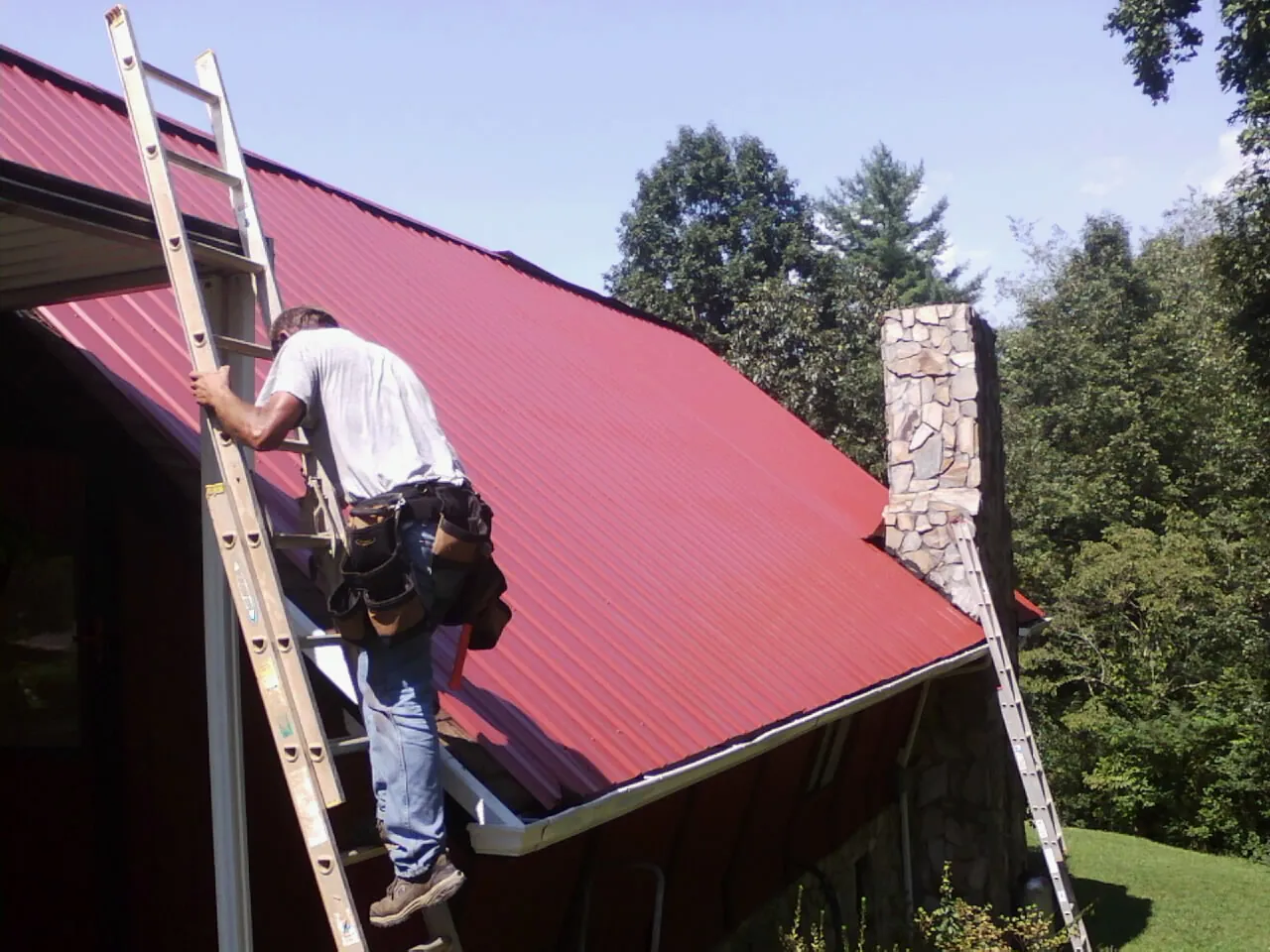 Roofer on a ladder beside a red metal roof and stone chimney — Soffit & Fascia Repair by Roofing Brothers in Weatogue